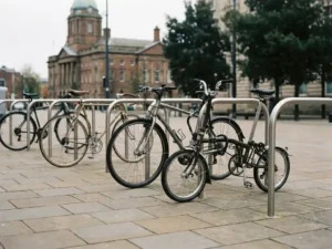Fahrradständer aus Stahl auf einem städtischen Platz mit geparkten Fahrrädern, Pflastersteinboden und Rathaus im Hintergrund.