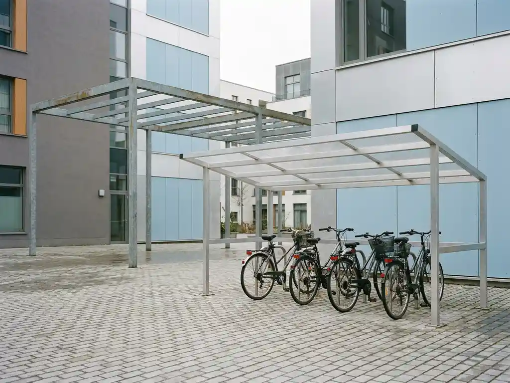 Aluminium-Fahrradunterstand mit transparentem Polycarbonat-Dach neben einem offenen Überdachungsystem auf einem Stadtplatz mit Kopfsteinpflaster.