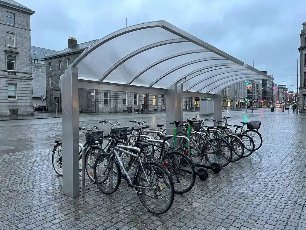 Aluminium-Fahrradüberdachung mit geschwungenem Polycarbonat-Dach auf urbanem Pflasterplatz, mehrere Fahrräder ordentlich abgestellt, nasses Kopfsteinpflaster.